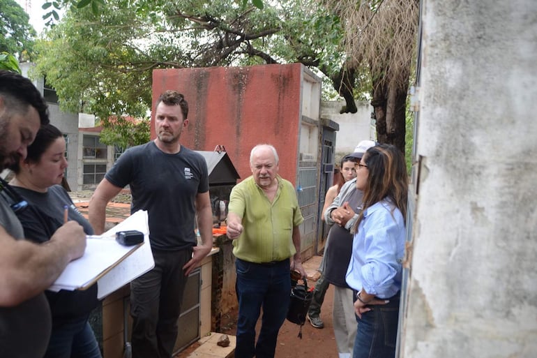 Grupo de seis personas en el cementerio del Este, discutiendo. Un hombre con cuaderno, mujeres atentas, ambiente serio y reflexivo.