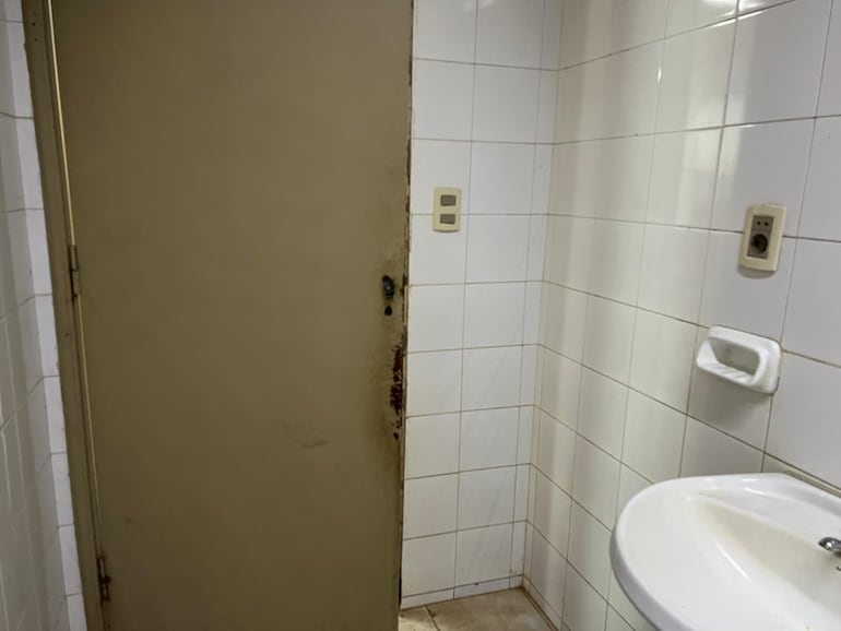 El sanitario se encuentra en condiciones precarias, con la puerta oxidada y sin picaporte.