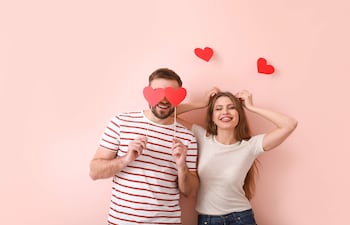 Hombre con barba en camiseta de rayas sosteniendo un corazón, junto a mujer con camiseta gris y corazones en la cabeza, ambos alegres.