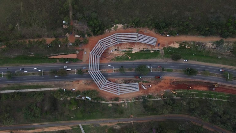 Vista aérea del viaducto en construcción, rodeado de vegetación y vehículos en la carretera, sin personas visibles.