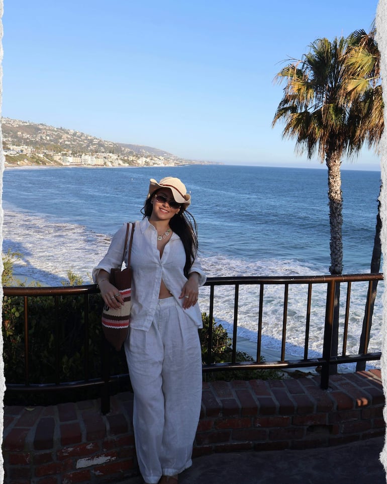 Mujer en conjunto blanco y sombrero claro sonríe frente al mar, rodeada de palmeras.
