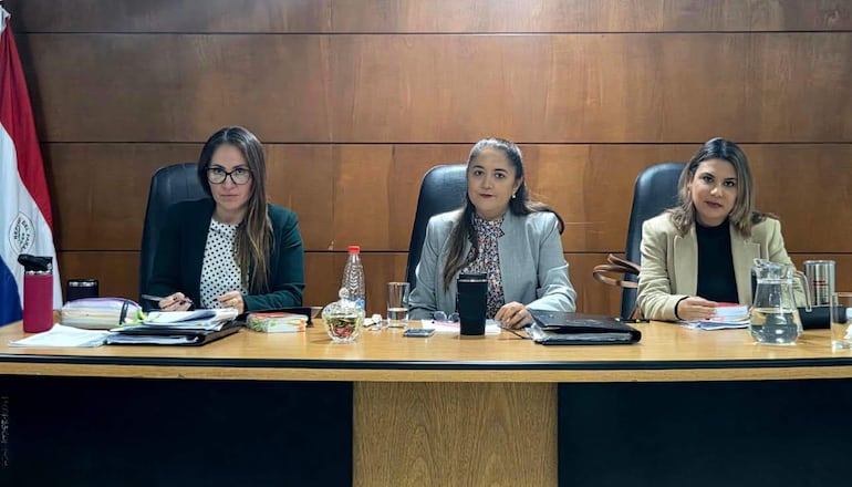 Cáceres con abrigo negro y gafas, Morel en gris claro y Rodríguez Brozón con blazer beige, en una reunión interior enfocadas en documentos.