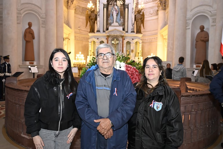 Josefina Torales con chaqueta negra, junto a un hombre canoso y una mujer con cinta nacional, en un entorno decorado florales.
