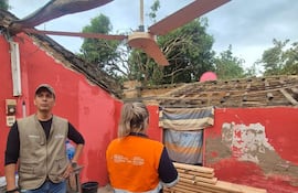 Hombre de pie con chaleco claro, escuchando a mujer de espaldas en camiseta naranja y azul, en ambiente deteriorado y húmedo.