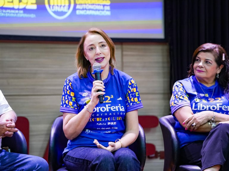 Mujer de cabello castaño claro con micrófono, dos personas observan atentamente; todos en camisetas azules que dicen 'LibroFeria'.