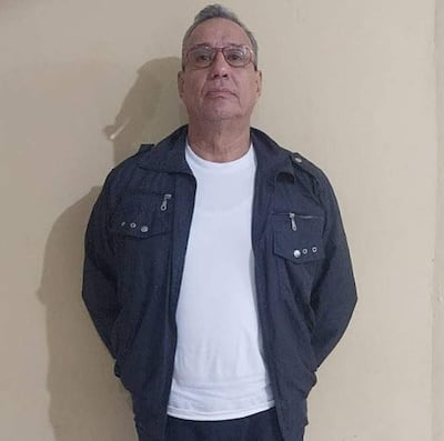 Hugo Javier González, con cabello gris y gafas, viste camiseta blanca y conjunto deportivo oscuro, de pie en una pared clara.