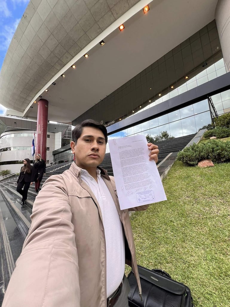 Hombre de pie con chaqueta clara y camiseta blanca sostiene un documento. Fondo de construcción moderna frente al Congreso.