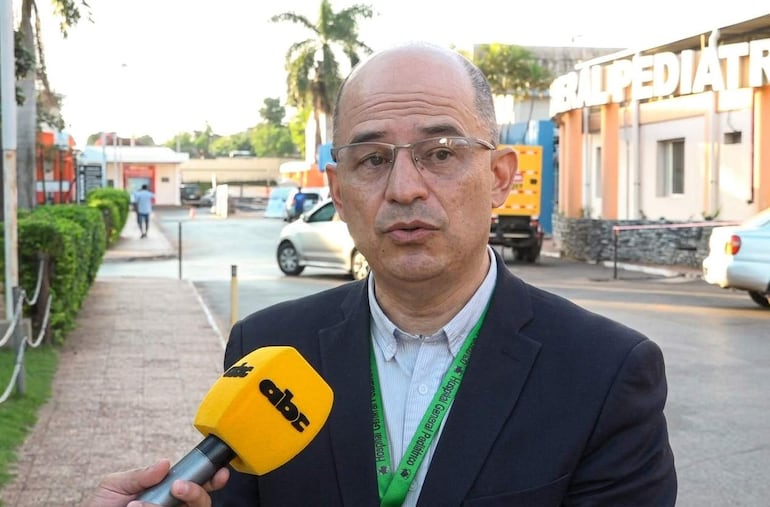 Dr. Héctor Castro en traje, sosteniendo un micrófono durante entrevista frente al Hospital General Pediátrico, fondo con algunas personas y vehículos.