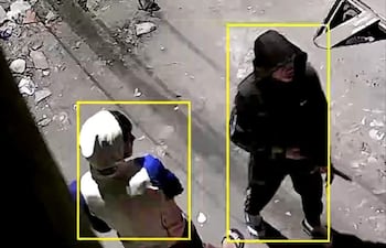 Dos hombres en la calle, uno con camiseta blanca y azul y gorro, otro con abrigo negro y un objeto que puede ser un arma.
