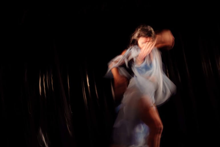 Mujer de cabello oscuro y vestimenta clara bailando en un escenario oscuro con atmósfera teatral.