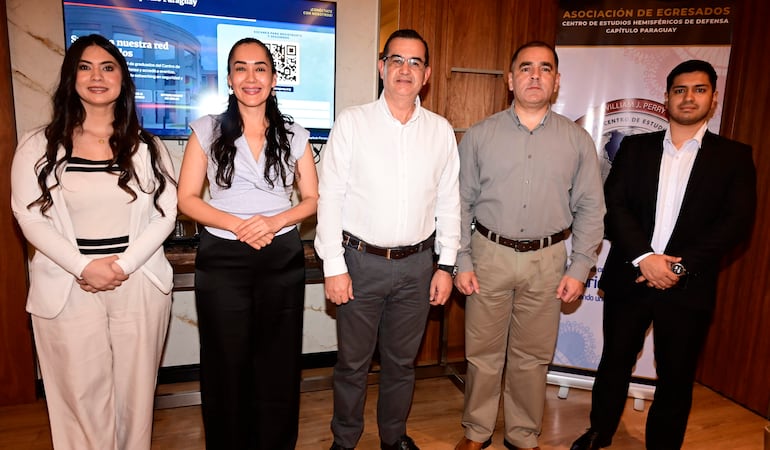 Cinco egresados sonríen, dos mujeres con blusas claras y tres hombres, uno en traje negro, en un ambiente iluminado con pantalla al fondo.