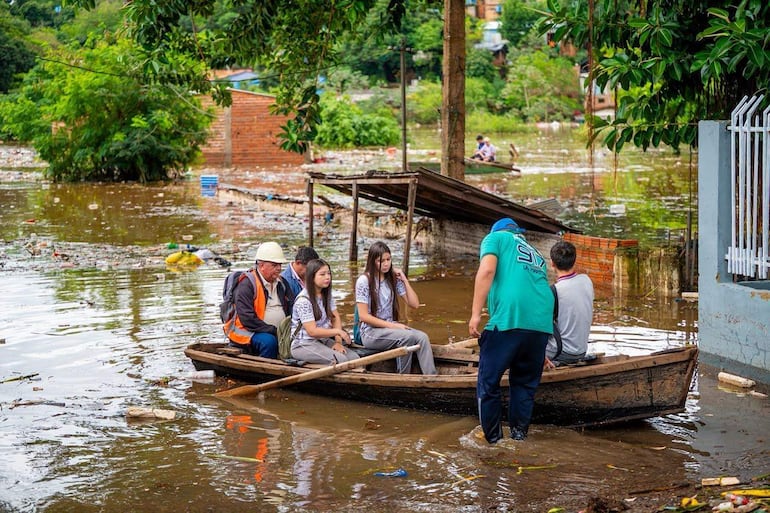 Hombre mayor con chaleco naranja y casco, junto a jóvenes en canoa, todos con expresiones de preocupación en un entorno inundado.