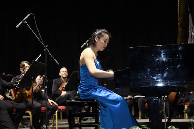 Pianista vestida de elegante azul se concentra frente a su piano; músicos de orquesta esperan detrás con ropa oscura.