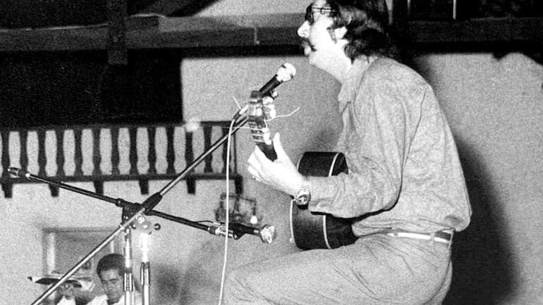 Hombre con cabello largo y gafas canta y toca guitarra en un escenario, concentrado en su actuación mientras un espectador lo observa.