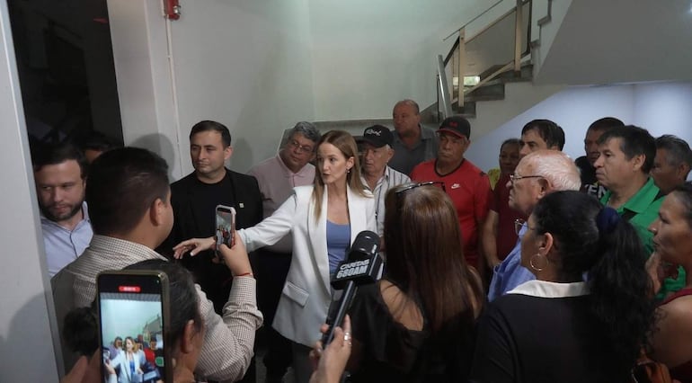 Soledad Núñez en prenda blanca habla a jubilados en gorras y camisetas de colores, en un ambiente cerrado lleno de teléfonos móviles.