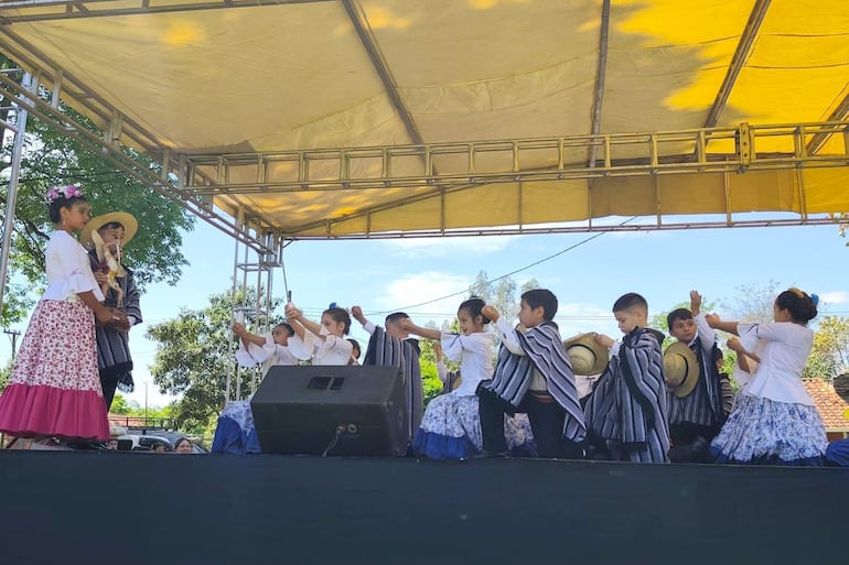11 niños en trajes típicos bailan en un escenario, bajo un toldo amarillo, con flores en el cabello y objetos en mano.