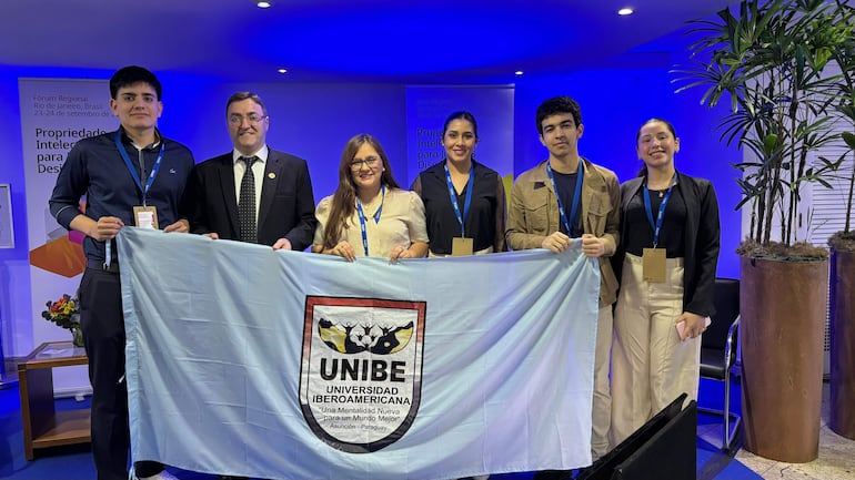 Grupo de seis personas sonriendo, entre ellas un hombre de traje negro, sosteniendo una bandera de UNIBE en un ambiente moderno con luces azules.