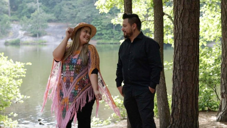Mujer con blusa colorida y sombrero sonríe junto a un hombre en camisa negra, en un entorno natural junto al lago.