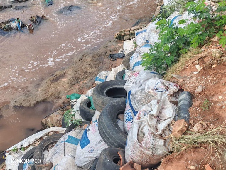 Entorno afectado por contaminación con neumáticos y sacos en la orilla de un arroyo marrón. Vegetación escasa.