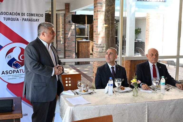 Yavuz Kül habla de pie en traje oscuro, mientras Ceferino Valdez y otro hombre lo escuchan sentados en una mesa decorada.