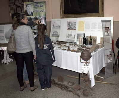 Mujer de pie explica a una niña con abrigo oscuro y detalles en rosa, rodeadas de objetos históricos en el Museo Simón Denis.