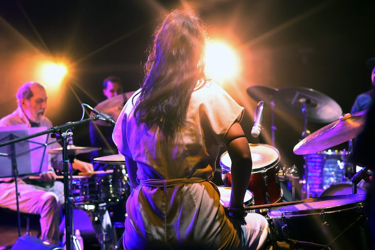 Mujer en blusa clara toca batería, mientras un hombre mayor con barba la acompaña en percusión, bajo luces brillantes.