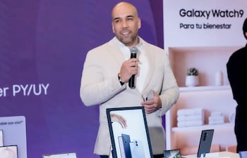 Hombre de pie con traje claro y celular, presenta productos Galaxy frente a mesa con dispositivos electrónicos y pantalla promocional.