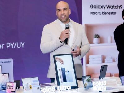 Hombre de pie con traje claro y celular, presenta productos Galaxy frente a mesa con dispositivos electrónicos y pantalla promocional.