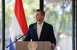 Santiago Peña Santi en podio, sonriendo y hablando, con saco oscuro y camisa clara, frente a la bandera de Paraguay.