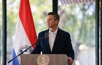 Santiago Peña Santi en podio, sonriendo y hablando, con saco oscuro y camisa clara, frente a la bandera de Paraguay.