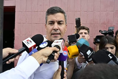 Santiago Peña con camisa blanca habla enérgicamente mientras una multitud de reporteros sostiene micrófonos, fondo rosa claro.