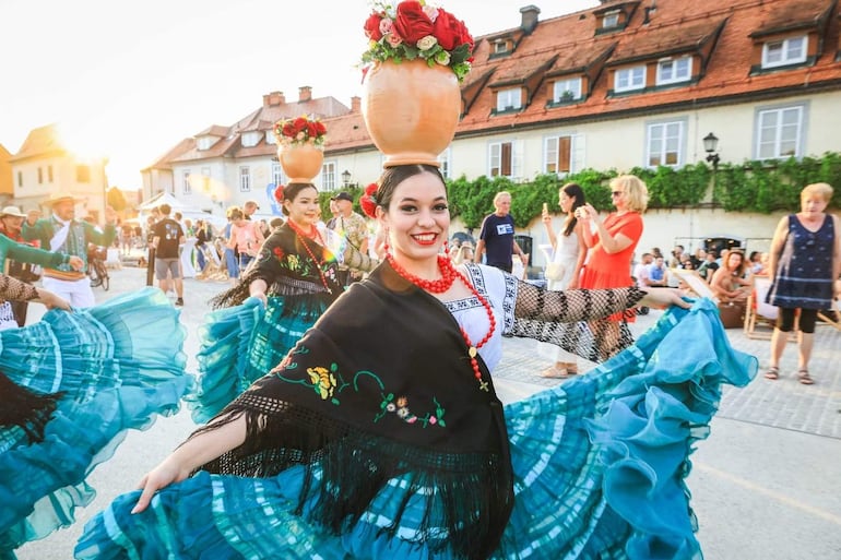 Bailarinas sonrientes en vestidos coloridos, con macetas decoradas en la cabeza, mientras bailan al sol con público observando.