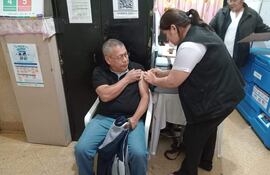 Hombre mayor en camiseta negra recibe vacuna del brazo izquierdo, asistido por mujer con camisa blanca y chaleco negro en centro de salud.
