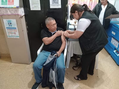 Hombre mayor en camiseta negra recibe vacuna del brazo izquierdo, asistido por mujer con camisa blanca y chaleco negro en centro de salud.