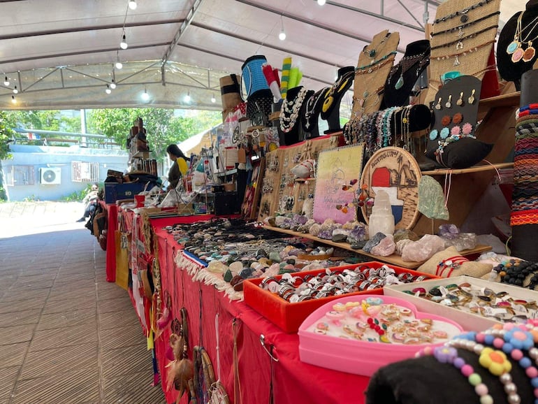 Mesa con artesanías coloridas sobre manteles rojos, rodeada de un ambiente urbano en un mercado al aire libre.