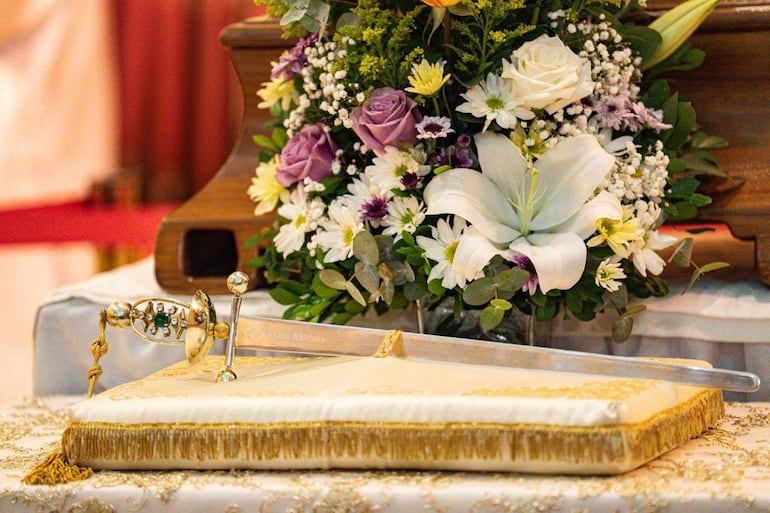 Espada decorada sobre cojín dorado rodeada de flores en ambiente ceremonial, en una mesa de la Catedral Metropolitana.