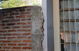 Pared con grietas en la Escuela Básica N°3770 San Miguel, rodeada de vegetación y con siluetas de personas en el fondo.