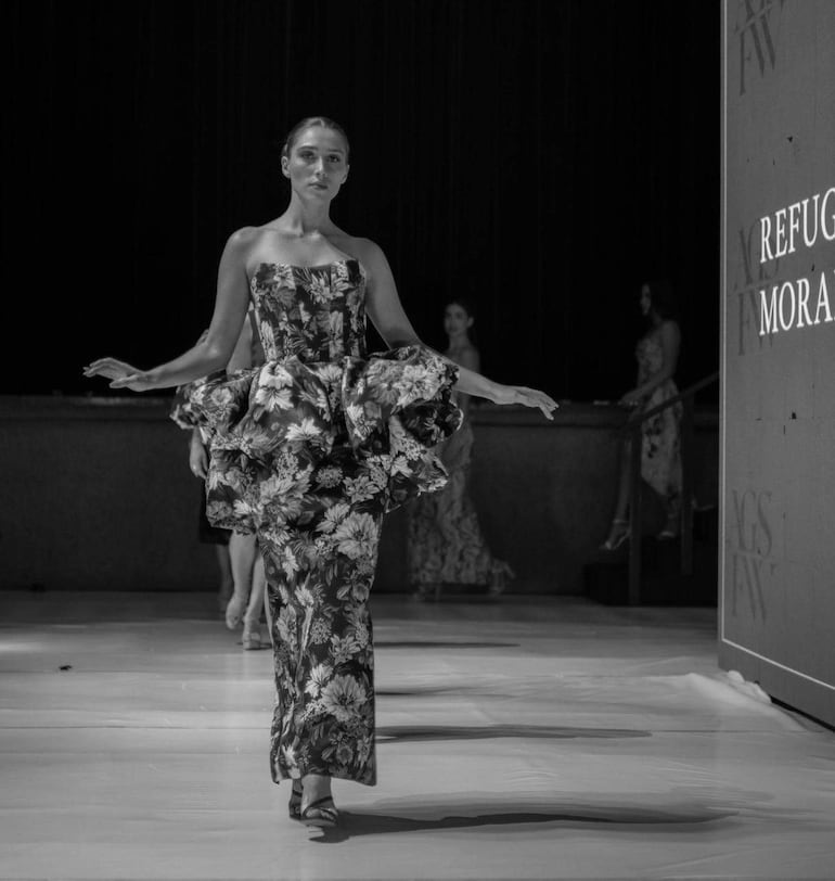 Modelo con vestido floral voluminoso camina con confianza en pasarela, con otras modelos en el fondo y pantalla que dice 'Refugio Moral'.