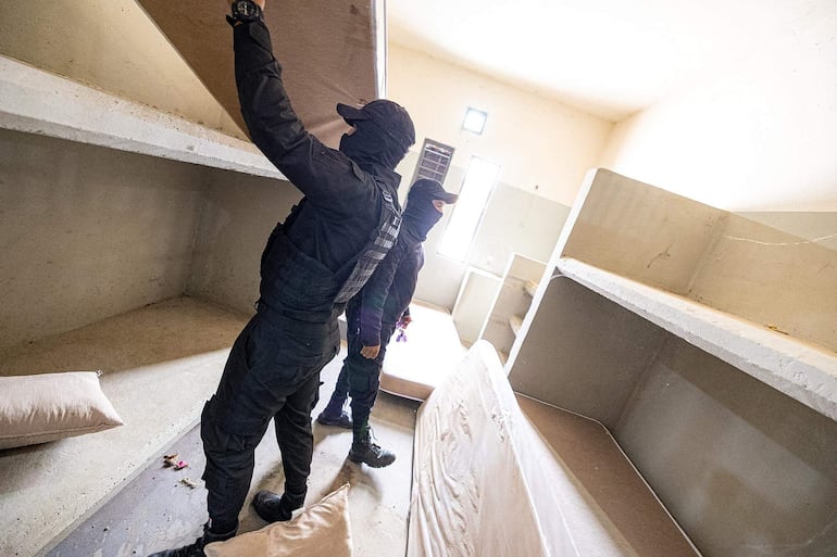 Dos hombres con uniformes oscuros y pasamontañas en un ambiente carcelario, levantando una tabla mientras observan el lugar.