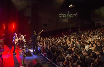 Tres músicos en escenario iluminado, interactuando con el público en un ambiente lleno de gente disfrutando el concierto.