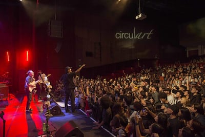 Tres músicos en escenario iluminado, interactuando con el público en un ambiente lleno de gente disfrutando el concierto.
