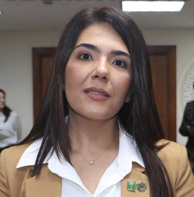 Persona con cabello largo en blusa blanca y sacón beige, conversando en un entorno formal con paredes de madera.