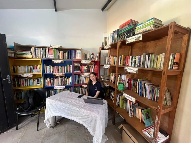 Mujer con cabello largo y oscuro sonríe en una mesa con computadora, rodeada de estanterías de libros en un ambiente interior.
