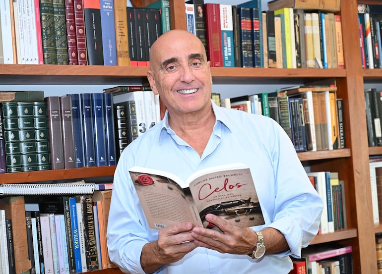 Hombre canoso sonriente en camisa clara sosteniendo el libro 'Celos', rodeado de estanterías de libros en un ambiente cálido.