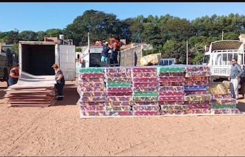 Seis personas en uniformes anaranjados y azules descargan cajas grandes en un terreno al aire libre, con camiones y árboles de fondo.
