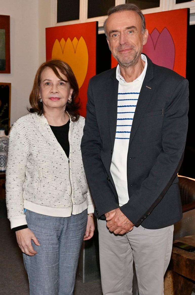 Mujer con chaqueta clara y hombre con blazer oscuro sonríen en un entorno decorativo con pinturas de flores.