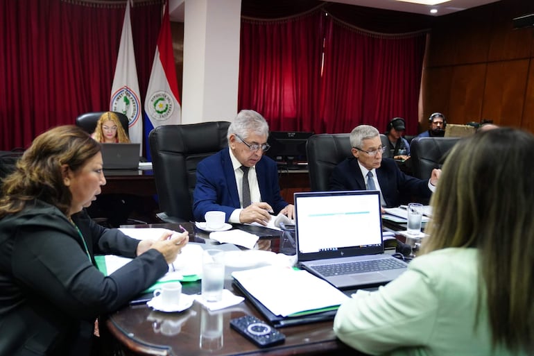 José Emilio Argaña Contreras escribe en la mesa rodeado de tres hombres y dos mujeres, en un ambiente formal con cortinas rojas.
