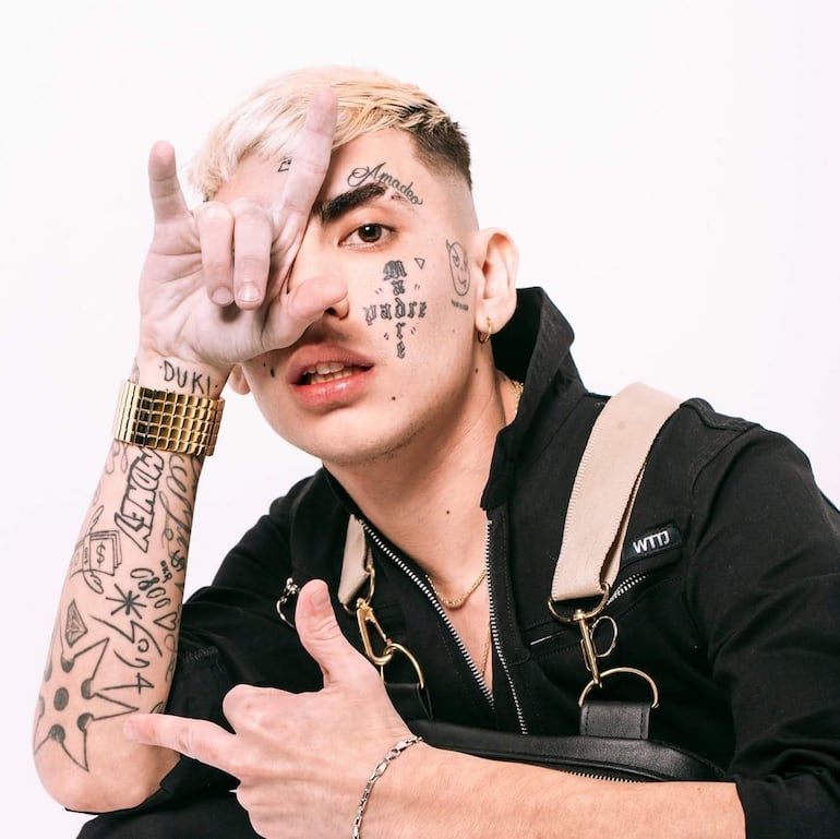 Joven de cabello rubio y tatuajes visibles, vestido con prenda negra, realizando un gesto de mano en fondo blanco.