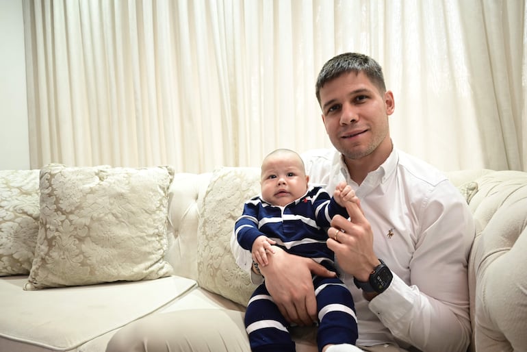 Charles Hockin, sonriente y en camisa blanca, sostiene a su bebé con ropa de rayas azul y blanca en un sofá con cortinas claras.