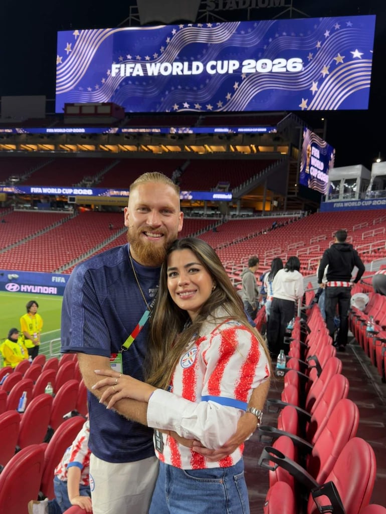 Isidro Pitta sonriente junto a mujer con camiseta de la selección paraguaya, en ambiente festivo dentro del estadio, con cartel de Mundial detrás.